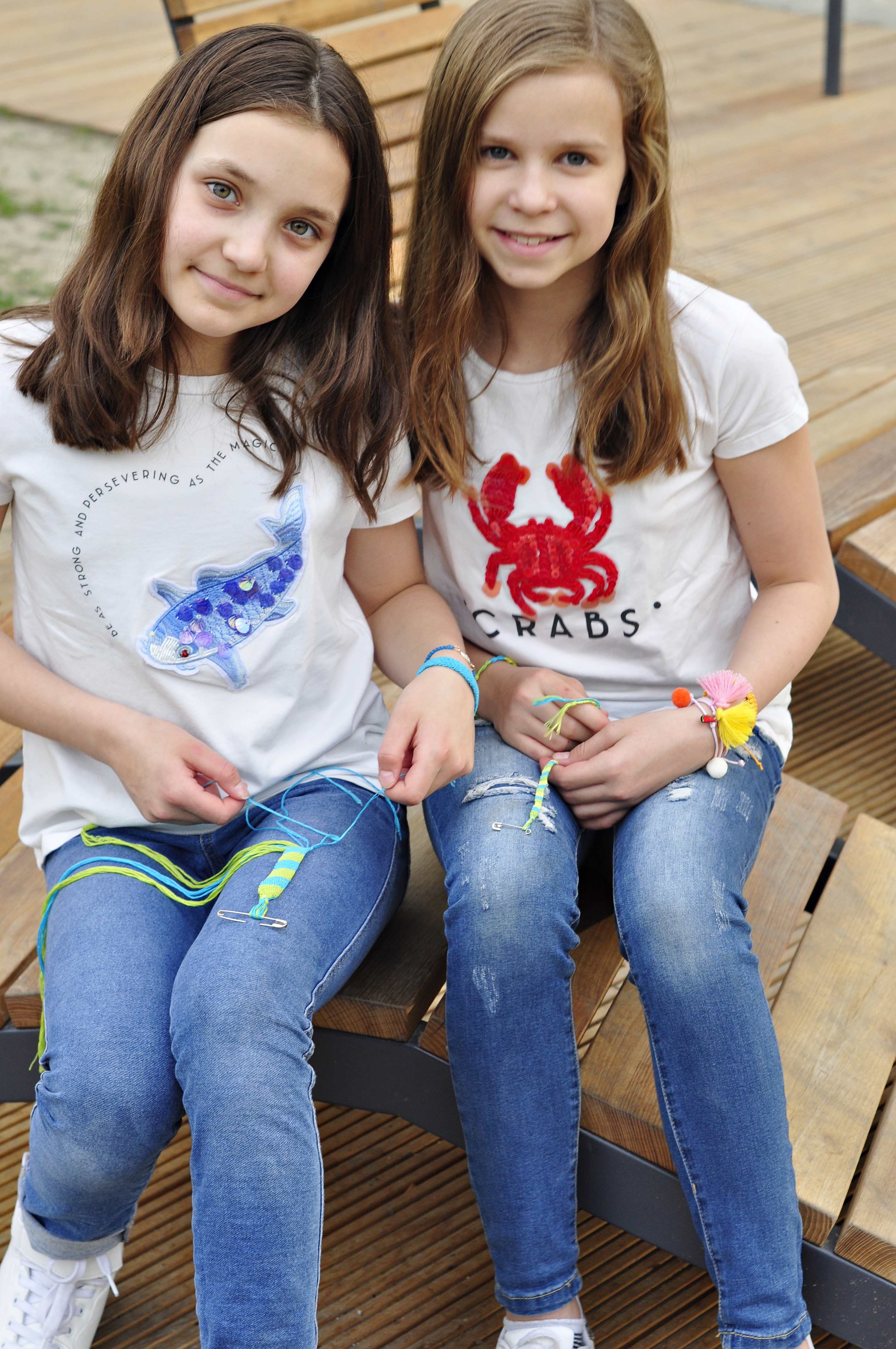 dziewczynki siedzą na ławce i pokazują jak zrobić bransoletkę z muliny